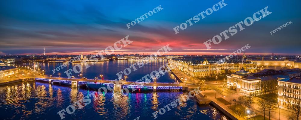 Панорамный ночной вид с дрона на Санкт-Петербург с Дворцовым мостом, рекой Невой, Адмиралтейством и Васильевским островом. Разведённый мост и огни вечернего города создают атмосферный городской пейзаж северной столицы России. Историческая архитектура, отражения подсветки в воде и масштабная панорама центра Петербурга подчёркивают красоту одного из самых известных городов России. Фотография идеально подходит для публикаций о путешествиях по России, мостах Санкт-Петербурга, ночной архитектуре, туристических маршрутах и аэрофотосъёмке города.